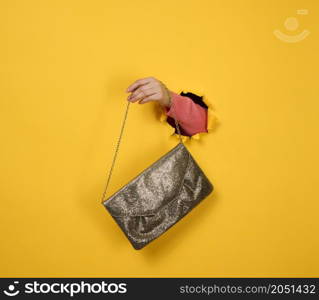 female hand is holding a small golden clutch bag with a metal chain on a yellow background. Part of the body sticking out of a torn hole in a paper background