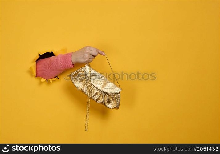 female hand is holding a small golden clutch bag with a metal chain on a yellow background. Part of the body sticking out of a torn hole in a paper background