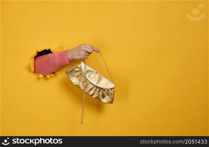 female hand is holding a small golden clutch bag with a metal chain on a yellow background. Part of the body sticking out of a torn hole in a paper background