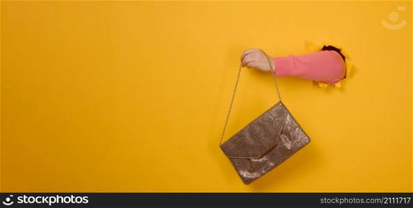 female hand is holding a small golden clutch bag with a metal chain on a yellow background. Part of the body sticking out of a torn hole in a paper background. Copy space