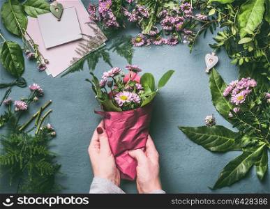 Female hand making beautiful bouquet of pink flowers on florist workspace, top view. Woman wrap bouquet in wrapping paper, step by step