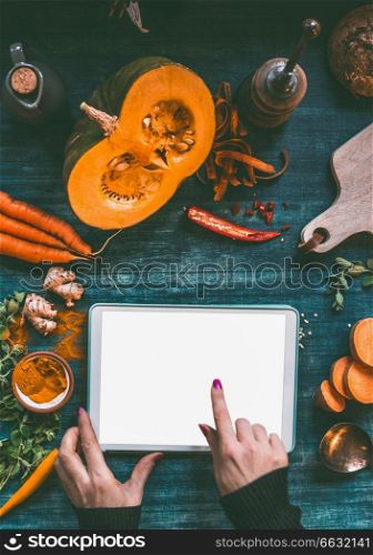 Female hand pointing with finger on tablet pc with mock up screen on dark kitchen table with orange cooking ingredients: pumpkin, sweet potato, carrots, turmeric powder,chili and ginger, top view