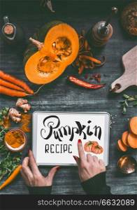 Female hand pointing with finger on tablet pc with text lettering  Pumpkin recipe on kitchen table background with orange cooking ingredients  sweet potato, carrots, turmeric powder,chili and ginger