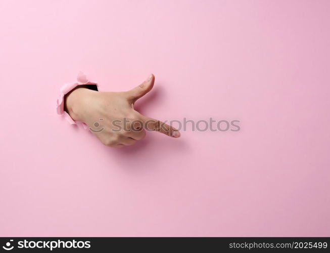 female hand sticks out of a torn hole in a pink paper background, part of the body points with the index finger to the side