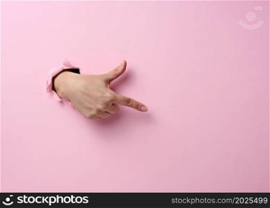 female hand sticks out of a torn hole in a pink paper background, part of the body points with the index finger to the side