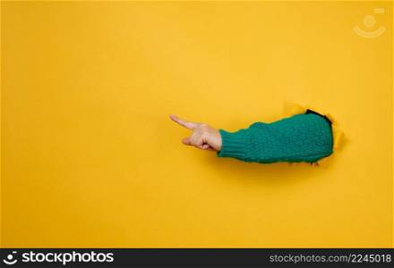 female hand sticks out of a torn hole in a yellow paper background, part of the body points with the index finger to the side. Copy space