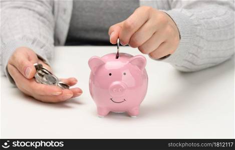 female hand throws a coin into a pink piggy bank on a white table. Concept of accumulating cash, saving, receiving subsidies