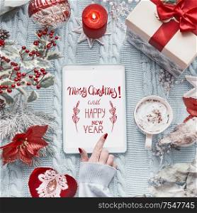Female hand touches tablet computer with Merry Christmas and Happy New Year greeting card on light blue knitted blanket with red Christmas decoration and burning candles. Top view. Flat lay