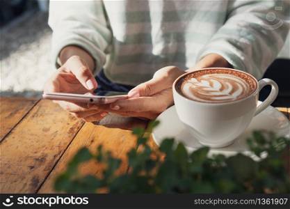 female hand using mobile phone with a cup of hot cocoa or chocolate on wooden table, close up