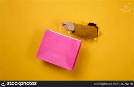 female hand with a pink paper shopping bag sticking out of a torn hole in a yellow paper background. Refusal from plastic, start of sales