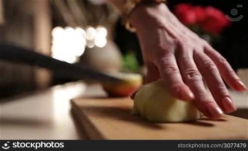 Female hand with knife slicing into pieces peeled apple on wooden chopping board in kitchen while preparing healthy fruits smoothie. Middle section of woman chopping apple on cutting board. Side view closeup.