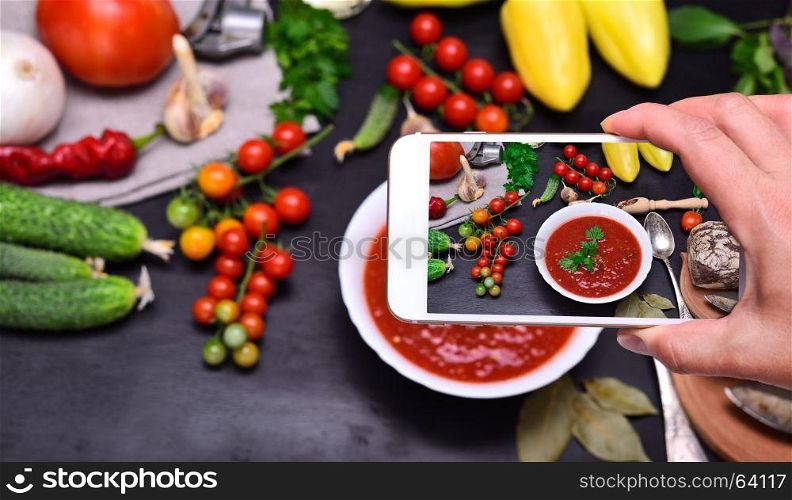 Female hand with smartphone in hand photographs soup gazpacho from red tomato in white round plate, top view