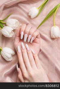 Female hand with spring nail design. White nail polish manicure. Female model hand with perfect manicure hold white tulip flowers on beige silk background