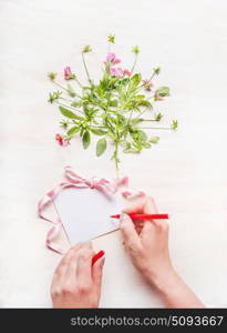 Female hand Write a greeting card on white wooden background with flowers bunch and pink ribbon, top view, copy space, mock up