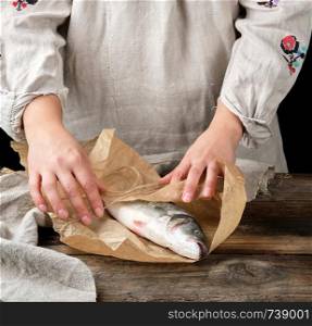 female hands are wrapped in brown paper whole fresh sea bass fish on a wooden table, black background