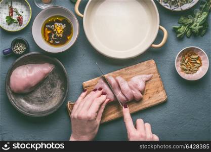 Female hands cut chicken breast on wooden cutting board on kitchen table background with ingredients, top view. Dieting cooking eating and healthy food concept