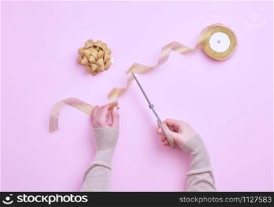 female hands cut golden ribbons in a roll for packing gifts, pink background, top view