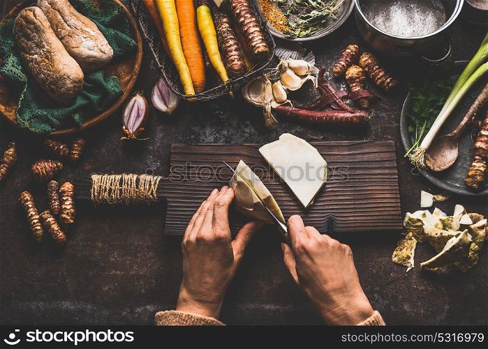 Female hands cutting celery on dark rustic kitchen table background with various vegetables and utensils. Root vegetables cooking preparation for tasty autumn dishes, top view. Healthy eating concept