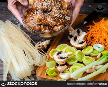 Female hands hold a bowl of chicken with teriyaki sauce and sesame seeds. Cooking noodles at home, close-up, dark wooden background, ingredients for cooking noodles.. Female hands hold a bowl of chicken with teriyaki sauce and sesame seeds.