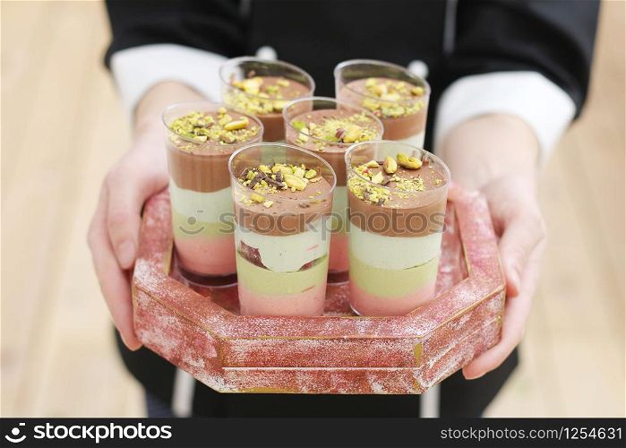 female hands hold a tray with six cups with a puff dessert, sprinkled with pistachios and chocolate on top.