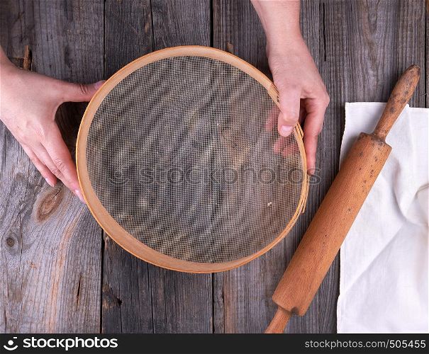 female hands holding a round wooden sieve for flour, next to an old wooden rolling pin on a gray table
