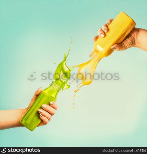 Female hands holding bottles with yellow and green splash smoothie or juice on blue background with tropical leaves and fruits. Summer beverages concept.