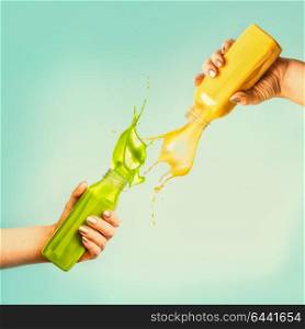 Female hands holding bottles with yellow and green splash smoothie or juice on blue background with tropical leaves and fruits. Summer beverages concept.