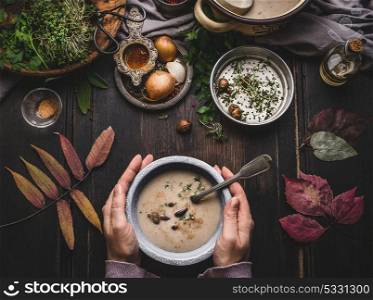 Female hands holding bowl with autumn vegetables chestnuts creamy soup on dark rustic kitchen table background with fall leaves, top view. Seasonal food and eating