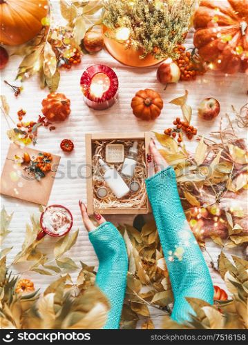 Female hands holding box with cosmetic products on desktop with autumn leaves, pumpkins,burning candles and cup of hot chocolate or coffee. Skincare routine for fall. Beauty box. Flat lay. Blog layout