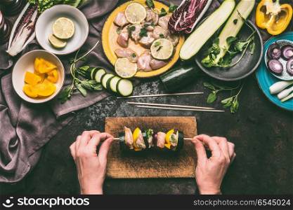 Female hands holding Chicken meat and vegetables skewer on cutting board on dark kitchen table background with plates and bowls ingredients, top view. Grill preparation