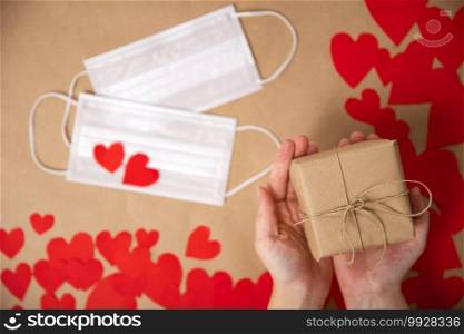 Female hands holding gift box for Valentines Day wrapped in Craft paper top view, romantic red hearts and Medical mask on background for Coronavirus, Covid-19 and Holliday, Valentine, Birthday concept. copy space. Female hands holding gift box for Valentines Day wrapped in Craft paper top view, romantic red hearts and Medical mask on background for Coronavirus, Covid-19 and Holliday, Valentine, Birthday concept.