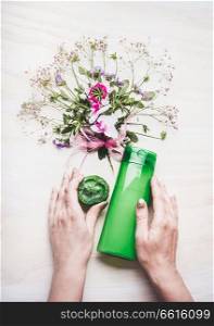 Female hands holding green cosmetic product bottle and handmade soap on white background with plants and flowers , top view. Natural cosmetic concept