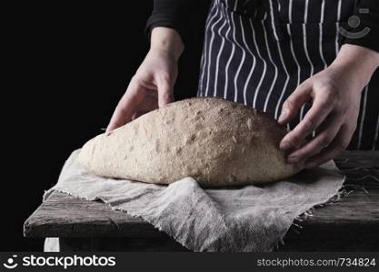 female hands holding oval baked wheat flour bread over wooden table, black background