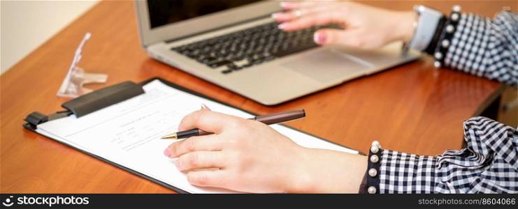 Female hands holding pen under the document and working with laptop at the table in an office. Female working with the documents