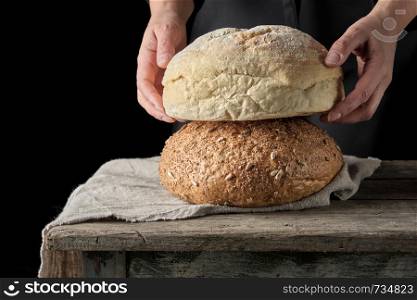 female hands holding round baked wheat flour bread over wooden table, black background