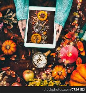Female hands holding tablet pc with pumpkins photo on dark background with cup of hot chocolate and autumn arrangement of fall leaves, burning candles, fall flowers, apples, fairy lights with bokeh