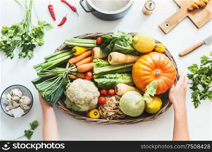 Female hands holding tray with various colorful organic farm vegetables on white desk background with pot , cutting board and knife. Healthy food and clean seasonal eating flat lay. Top view.