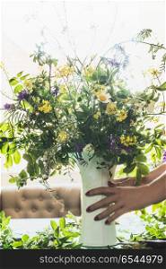 Female hands holding vase with summer bouquet made from field wild flowers on a table in modern living room at window. Summer still life. Cozy home scene
