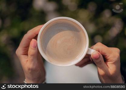Female hands holding white porcelain cup of coffee. Fresh hot drink with foam from coffee-machine. High quality photo. Female hands holding white porcelain cup of coffee. Top view. Fresh hot drink with foam from coffee-machine. Caffeine, energy, aroma concept
