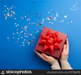 female hands holds a beautiful shiny red box with a big red bow on a blue background with multi-colored sparkles, concept of congratulations and surprise, top view