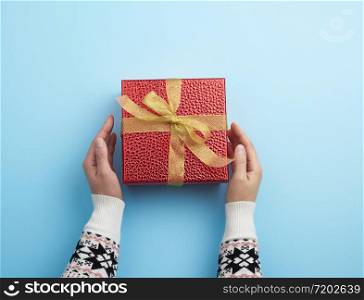 female hands holds a red box tied with a golden ribbon, concept of congratulations and gifts, top view, blue background