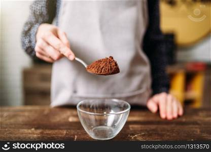 Female hands holds spoon with chocolate powder over glass bowl. Sweet cake cooking preparation