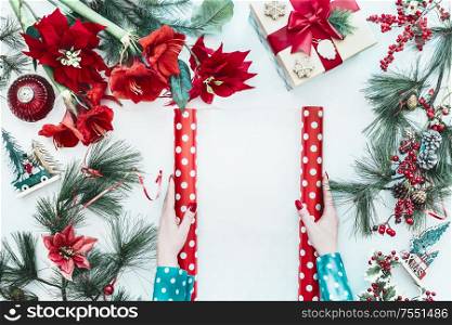 Female hands in blue blouse holding red polka dot wrapping paper with copy space on white desk with Christmas flowers , gift box and fir branches, top view. Flat lay