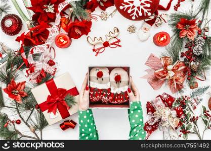 Female hands in green blouse holding open gift box with funny deer socks on white background with red Christmas decoration with fir branches and candles. Top view. Flat lay