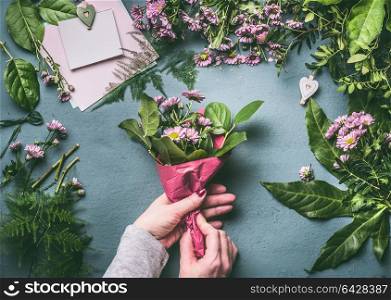 Female hands making lovely bouquet of pink flowers with wrapping paper on florist workspace, top view. Florist work, step by step