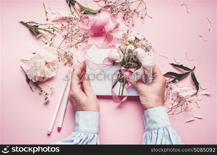 Female hands making lovely floral arrangement with flowers and envelop on pale pink background, top view. Creative greeting, Invitation and holiday concept