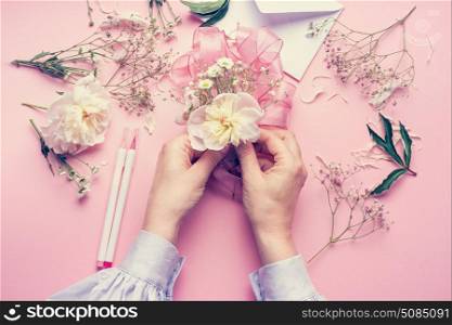 Female hands making lovely floral arrangement with flowers and ribbon on pale pink background, top view. Creative greeting, Invitation and holiday concept