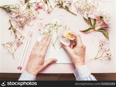 Female hands making lovely floral arrangement with flowers in opened envelop on pale pink background, top view. Creative greeting, Invitation and holiday concept