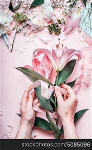 Female hands making lovely pink lily flowers bouquet on pastel table with florist decoration equipment, top view. Creative Florist workspace and flowers arrangement. Festive holiday concept.