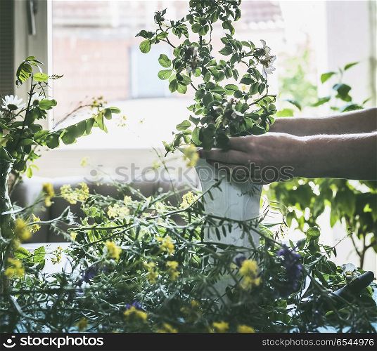Female hands making summer bouquet with wild flowers on a table in modern living room at window. Summer still life. Cozy home scene